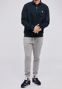 Marinblå zip-up sweatshirt med en liten logotyp, grå sweatpants och marinblå sneakers med röda detaljer. Avslappnad, atletisk outfit, mjukt tygtextur.