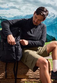Sac à dos en tissu noir avec plusieurs poches, homme portant une polaire noire zippée et un short vert olive, assis sur une souche dans une zone montagneuse.