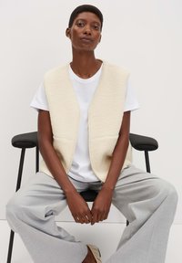 Offwhite Fleece-Weste mit weitem Halsauschnitt und vorderen Taschen, kombiniert mit einem weißen T-Shirt und hellgrauen, weit geschnittenen Hosen.