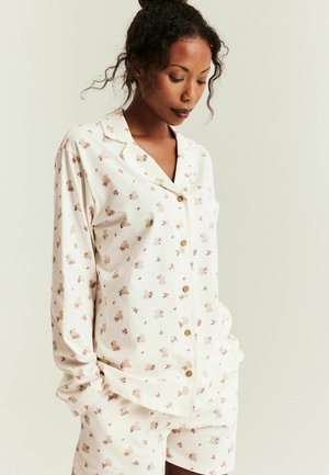 Vrouw draagt een crèmekleurig overhemd met knopen en bijpassende korte broek met kleine patronen van teddyberen en bloemen, staand met handen in de zakken.