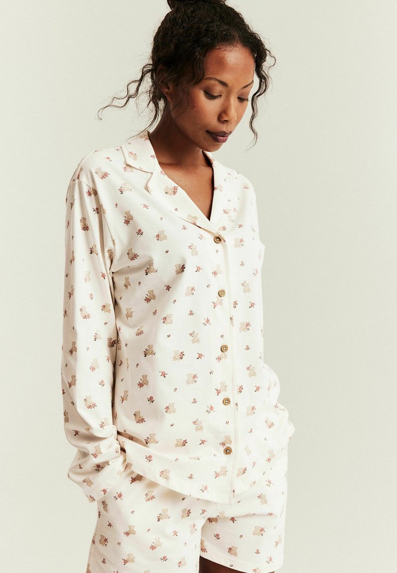 Frau trägt cremefarbenes Hemd mit Knöpfen und passende Shorts mit kleinen Teddybär- und Blumenmustern, steht mit den Händen in den Taschen.