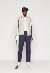 Persoon met een marineblauwe pet, een beige doorgestikte bodywarmer, een wit shirt met lange mouwen, marineblauwe broek en witte schoenen, die een golfclub vasthoudt met een handschoen aan de hand.