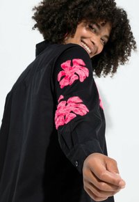 Schwarzes T-Shirt mit kurzen, strukturierten Ärmeln und texturierten pinken Blumenstickereien an beiden Armen. Glattes Material mit lockerer Passform.
