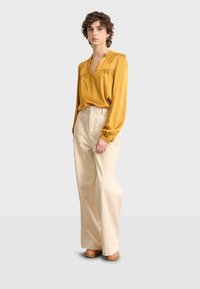 Mujer con cabello corto y rizado que lleva una blusa de manga larga mostaza, pantalones beige de pierna ancha y zapatos marrones con tachuelas, de pie contra un fondo claro.