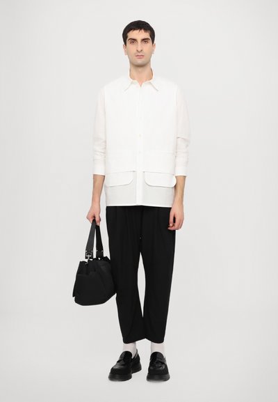 White button-up shirt, two front pockets, paired with loose black trousers and black shoes. Black bag in hand, with light socks visible.