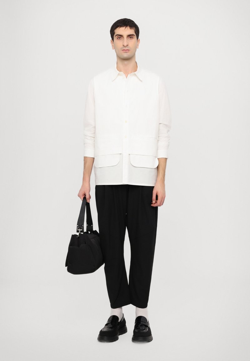 Camisa blanca de botones, con dos bolsillos frontales, combinada con pantalones negros holgados y zapatos negros. Bolso negro en la mano, con calcetines claros visibles.