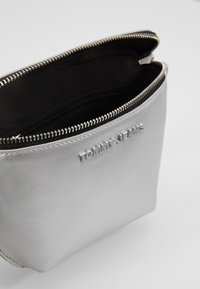 Pochette in pelle sintetica argento con superficie testurizzata, chiusura superiore con zip, fodera nera e logo "TOMMY JEANS" in rilievo sul fronte.