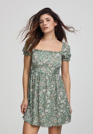 Abito floreale verde con scollo quadrato, maniche a palloncino corte e vita stretta. Il materiale in cotone presenta piccoli fiori rosa e bianchi.