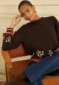 Ciemnobrązowy sweter dzianinowy z akcentami w panterkę, prążkowany dół i rękawy z mankietami, ozdobione czerwonymi i kremowymi paskami. Stylizowany z niebieskimi dżinsami.