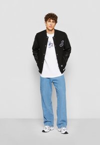 Sort varsity-jakke med læderærmer, bogstavet "S" på brystet, hvid t-shirt med grafisk print, lyseblå jeans og sneakers.