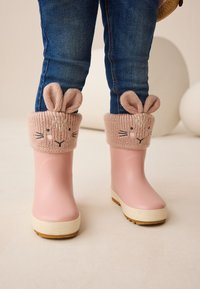 Rosa Gummistiefel mit einer cremefarbenen Sohle, versehen mit gestrickten, pastellrosa Bündchen, die Hasenohren und bestickte Gesichtszüge aufweisen.