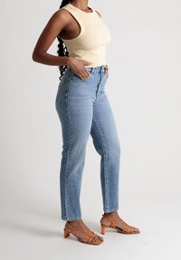 Vaaleankeltainen ribattu hihaton paita, korkeavyötäröiset siniset farkut ja oranssit remmillä varustetut korot. Sileä rakenne, istuva muotoilu, rento tyyli.