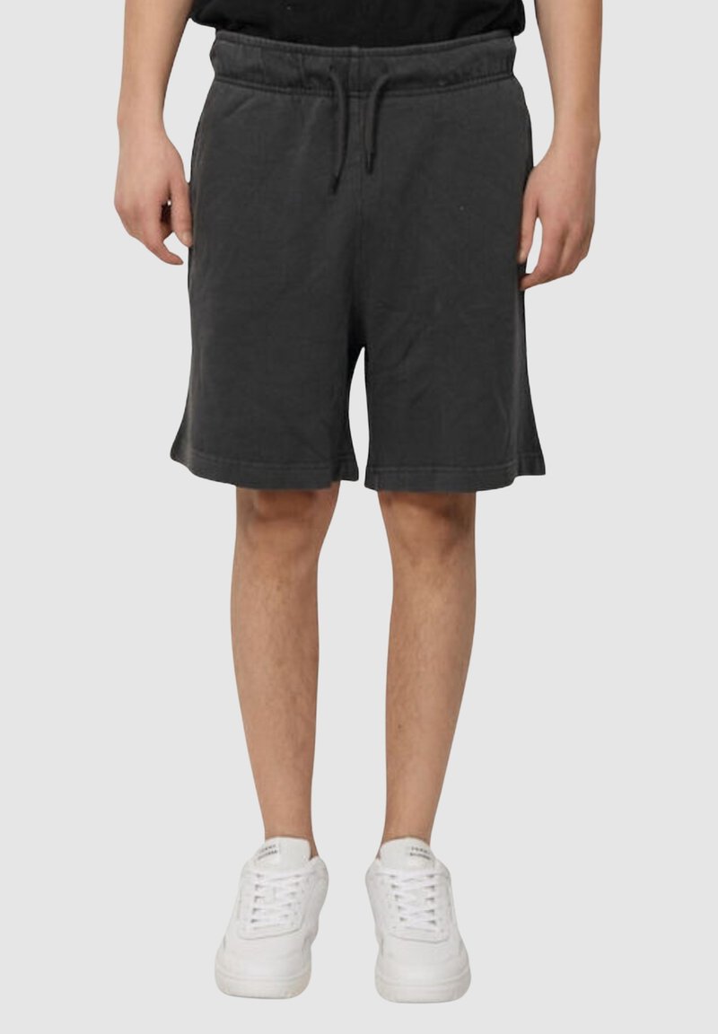 Person in schwarzen Shorts mit Kordelzug und weißen Turnschuhen, stehend vor einem schlichten hellgrauen Hintergrund.