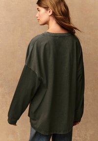 Chemise à manches longues, de couleur vert foncé, fabriquée en tissu doux. Présente une coupe décontractée, un col rond et des détails de couture le long des coutures.