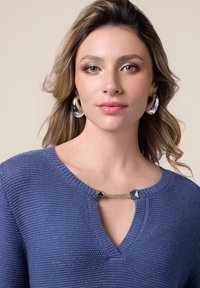 Pulover tricotat albastru cu decolteu în V și mâneci scurte, având o chaină argintie cu accente sub formă de piramidă la gât. Textură fină. Cercei cu inel argintiu.