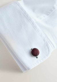 Weißes Hemdmanchet mit einem silbernen Knopf, der ein strukturiertes kastanienbraunes Muster aufweist. Glattes Gewebe, ordentlich gefalteter Rand und kontrastierende Nähte.