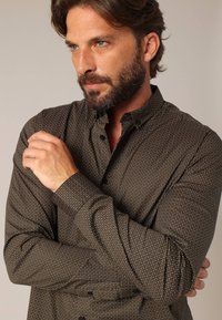 Bruine blouse met kleine witte stippen, met een gestructureerde kraag en lange mouwen. Gladde textuur en getailleerd ontwerp.