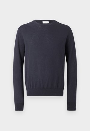 Mladý muž s kučeravými vlasmi, oblečený v čiernom svetri, svetlomodrých širokých džínsoch a čiernych topánkach, stojaci s jednou rukou v vrecku.