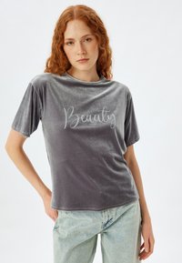 Camiseta de terciopelo gris con mangas cortas que presenta la palabra "Belleza" en script plateado. La tela tiene una textura suave y un ajuste ligeramente holgado.