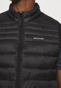 Schwarze Steppweste mit hohem Kragen, horizontalem Steppmuster, Frontreißverschluss und einem Markenpatch auf der linken Brustseite.