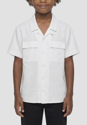 Bambino che indossa una camicia bianca a maniche corte con due tasche anteriori con patta e pantaloni neri, in piedi davanti a uno sfondo uniforme.