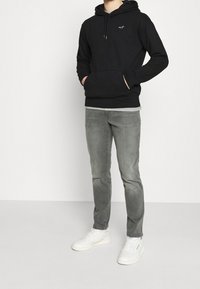 Hombre con sudadera negra, jeans grises y zapatillas blancas, de pie con las manos en los bolsillos de la sudadera, contra un fondo liso.