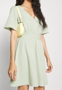 Světle zelené lněné šaty s výstřihem do V, krátkými rukávy, detaily na knoflíky a přiléhavým pasem. Kombinováno s malou žlutou taškou.