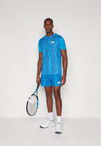 Sininen urheiluasu, joka koostuu lyhythihaisesta kuvioidusta paidasta ja sitä vastaavista shortseista. Asusteina tennismaila ja valkoiset lenkkarit.