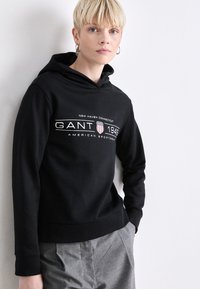 Svart hettegenser med en hvit logo som leser "GANT 1949" og teksten "NEW HAVEN CONNECTICUT AMERICAN SPORTSWEAR" på forsiden.