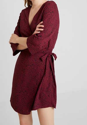 Vrouw draagt een bordeaux wikkeljurk met zwarte luipaardprint, driekwart mouwen en een strik in de taille, staat met gekruiste armen.