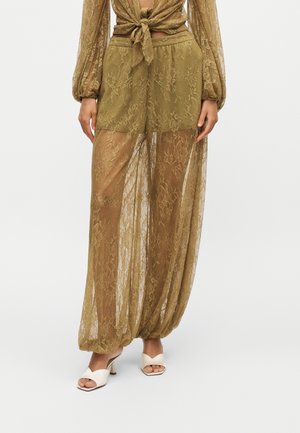 Indossa pantaloni trasparenti in pizzo verde oliva con shorts interni, abbinati a un top in pizzo coordinato legato in vita e sandali bianchi con tacco e punta aperta.