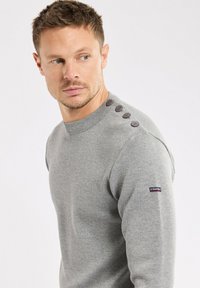 Maglione grigio a maglia con scollatura a girocollo, caratterizzato da una finitura testurizzata e quattro bottoni marroni sulla spalla. Toppa con logo sulla manica.