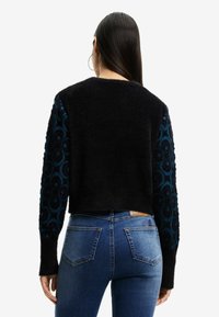 Svart kortärmad tröja med en texturerad, fluffig kropp och blå mönstrade ärmar. Parat med medelblå denimjeans med bakfickor.