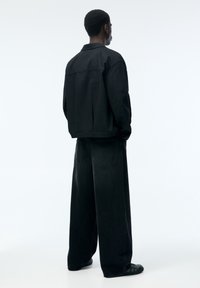 Musta oversize-farkkutakki korkealla kauluksella, pitkät hihat, yhdistetty leveälahkeisiin mustiin farkkuihin. Minimalistiset saumat, rento istuvuus, mattapintainen.