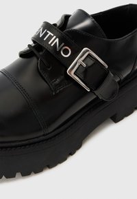 Sorte lærskos med patentfinish, spenne og sølvlogo. Tykk svart såle med en røff tekstur, minimalistisk design.