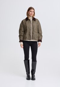 Olívazöld rövidített dzseki barna kordbársony gallérral, elöl patentos záródással és két ferde zsebbel. Fekete farmernadrág és térdig érő fekete csizmák.