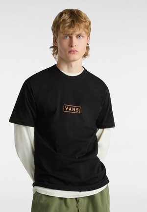 Czarna bawełniana koszulka z prostokątnym pomarańczowym logo "VANS" na piersi, nałożona na białą koszulę z długim rękawem, w połączeniu z zielonymi spodniami.