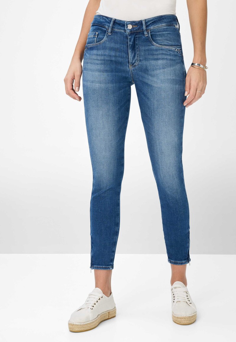 Denim jeans in medium blue wash with a slim fit, featuring a button closure, five pockets, and raw hem detailing at the ankle.