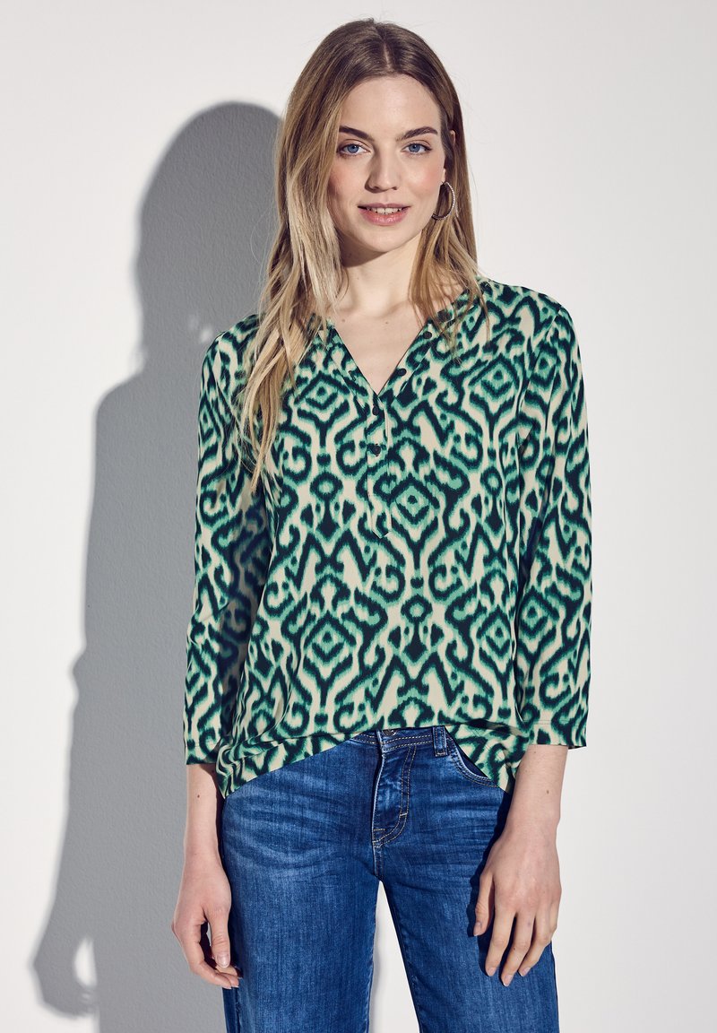 Blouse à motifs vert et crème avec un décolleté en V et des manches trois-quarts, associée à un jean en denim bleu. Tissu lisse et léger.
