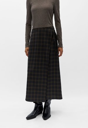 Femme portant une longue jupe portefeuille à carreaux foncés, un haut gris à manches longues et des bottines noires à bout pointu, debout sur fond blanc.