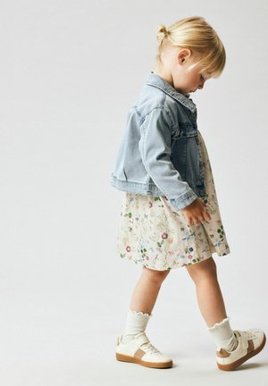 Mlad otrok z blond lasmi v figi, oblečen v cvetlično obleko, svetlo modro jeans jakno, bele nogavice in bež superge, hodi naprej.