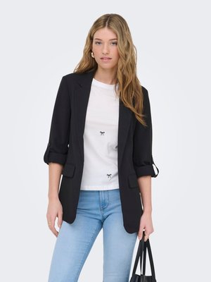 Junge Frau mit langen blonden Haaren, die einen schwarzen Blazer, ein weißes T-Shirt mit kleinen Schleifendrucken und hellblaue Jeans trägt und eine schwarze Handtasche hält.