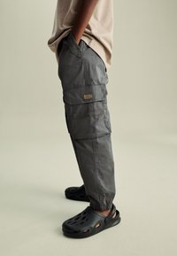 Pantaloni cargo grigi con polsini elasticizzati, numerose tasche e un'etichetta sul retro, abbinati a zoccoli neri ventilati.
