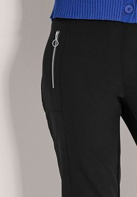 Zwarte broek met een strak design, voorzien van een zilveren ritszak aan de zijkant en een gladde textuur. Blauwe geribde trui zichtbaar erboven.