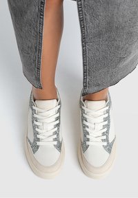 Sneakers bianchi con dettagli in glitter grigio, realizzati in materiali in suede testurizzato e pelle liscia, con lacci piatti e un design a punta arrotondata.