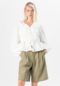 Valkoinen pallohaalarihiha-blouse, jossa on kerätty vyötärö ja etusolmu. Yhdistetty oliivinvihreisiin polvipitkiin taitetuilla lahkeilla shortsit.