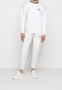 Valkoinen puuvillapaita, jossa on musta ribbauskaulus, ja siinä on kultainen brodeerattu logo. Yhteensopivat valkoiset puuvillajoggerit, joissa on samanlainen logo.