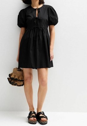 Robe noire à manches bouffantes avec un col à nouer, une taille cintrée et un ourlet arrivant au genou. Les accessoires comprennent un sac tressé et des sandales épaisses.