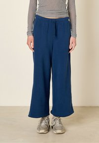 Pantalons de survêtement large bleu marine avec une taille élastique et un cordon de serrage, présentant une texture lisse. Portés avec des chaussures de sport argentées.