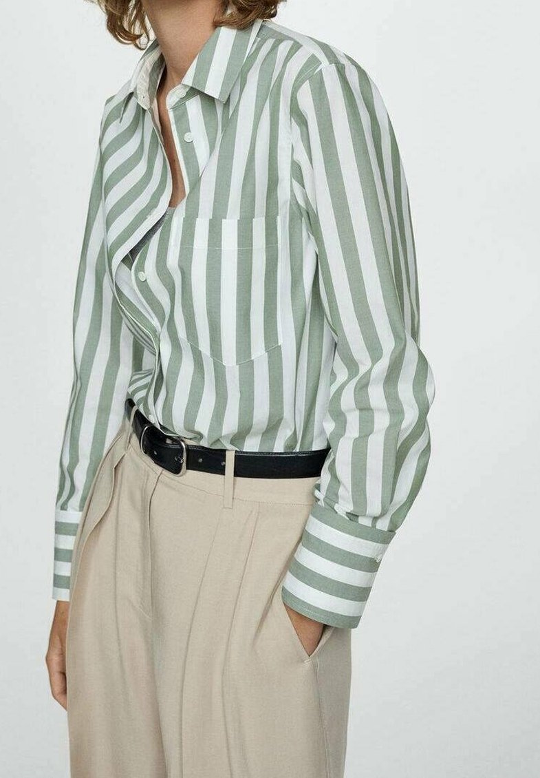 Personne portant une chemise à rayures verticales vertes et blanches rentrée dans un pantalon beige taille haute avec une ceinture noire, une main dans la poche.